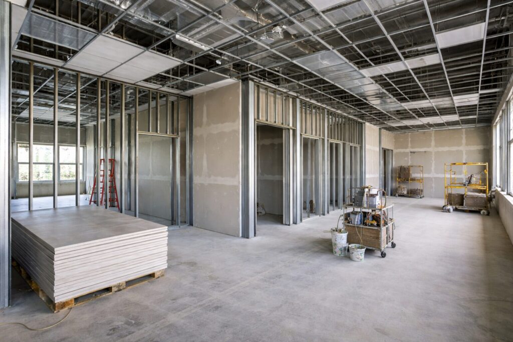 Interior commercial space under construction with metal stud framing, drywall installation, and exposed ceiling grid in a clean, organized job site